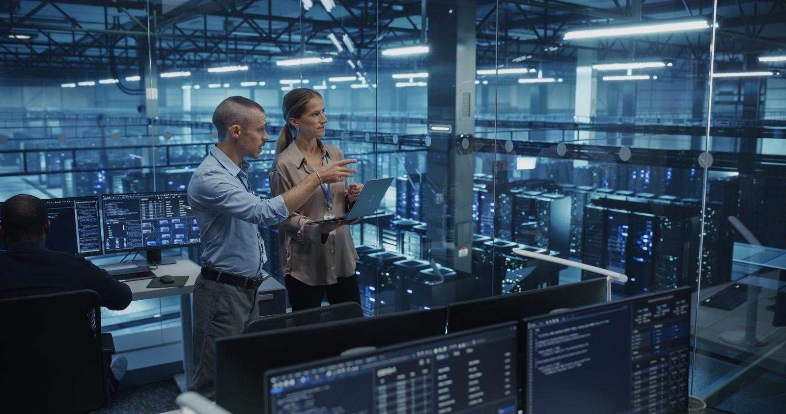 Two people with a laptop looking over rows of server racks through a glass wall