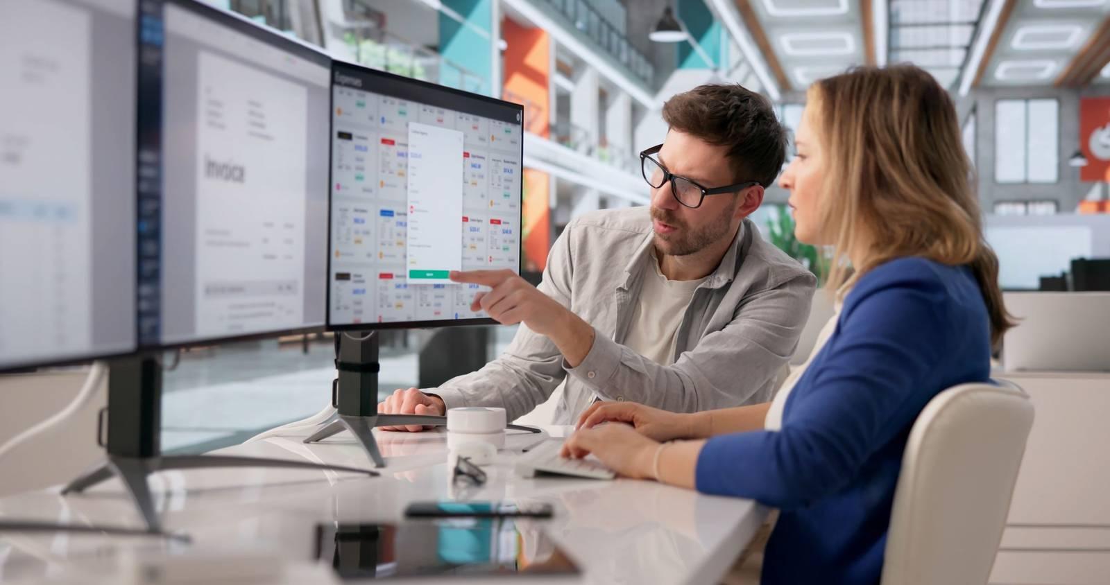 Two colleagues at a desk reviewing figures on a large computer monitor